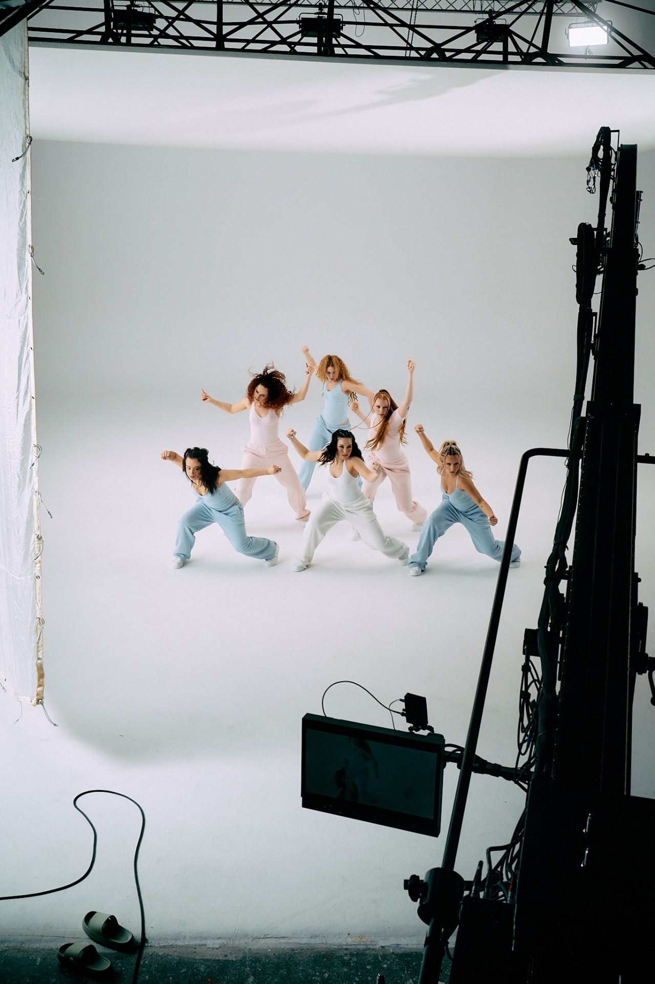 Behind the scenes — wide shot of the cast performing on a white cyclorama with lighting truss and monitor visible